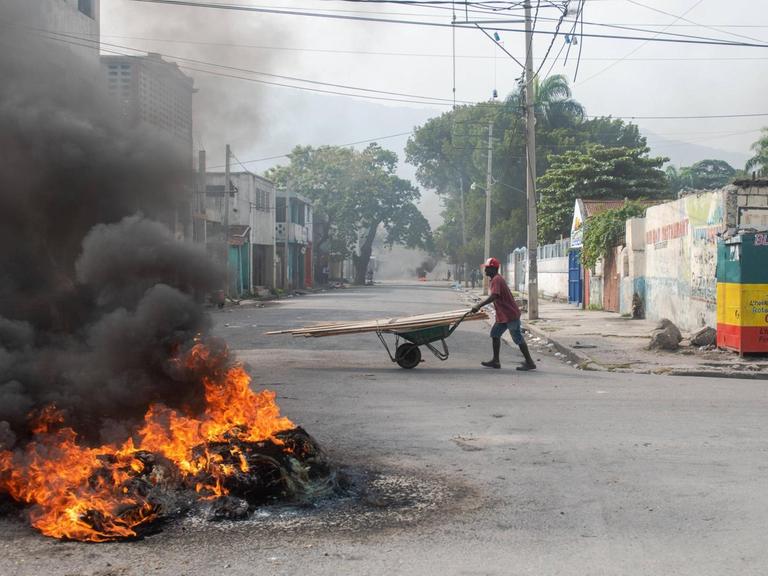 Eine brennende Barrikade in Haitis Hauptstadt Port-au-Prince. Dahinter geht eine Person mit einer Schubkarre die Straße entlang.