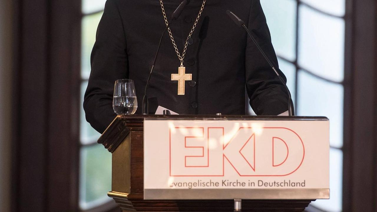 Ohne Kopf abgebildet steht ein Geistlicher mit einer Kette mit Kreuz hinter einem Rednerpult mit dem Schriftzug: EKD