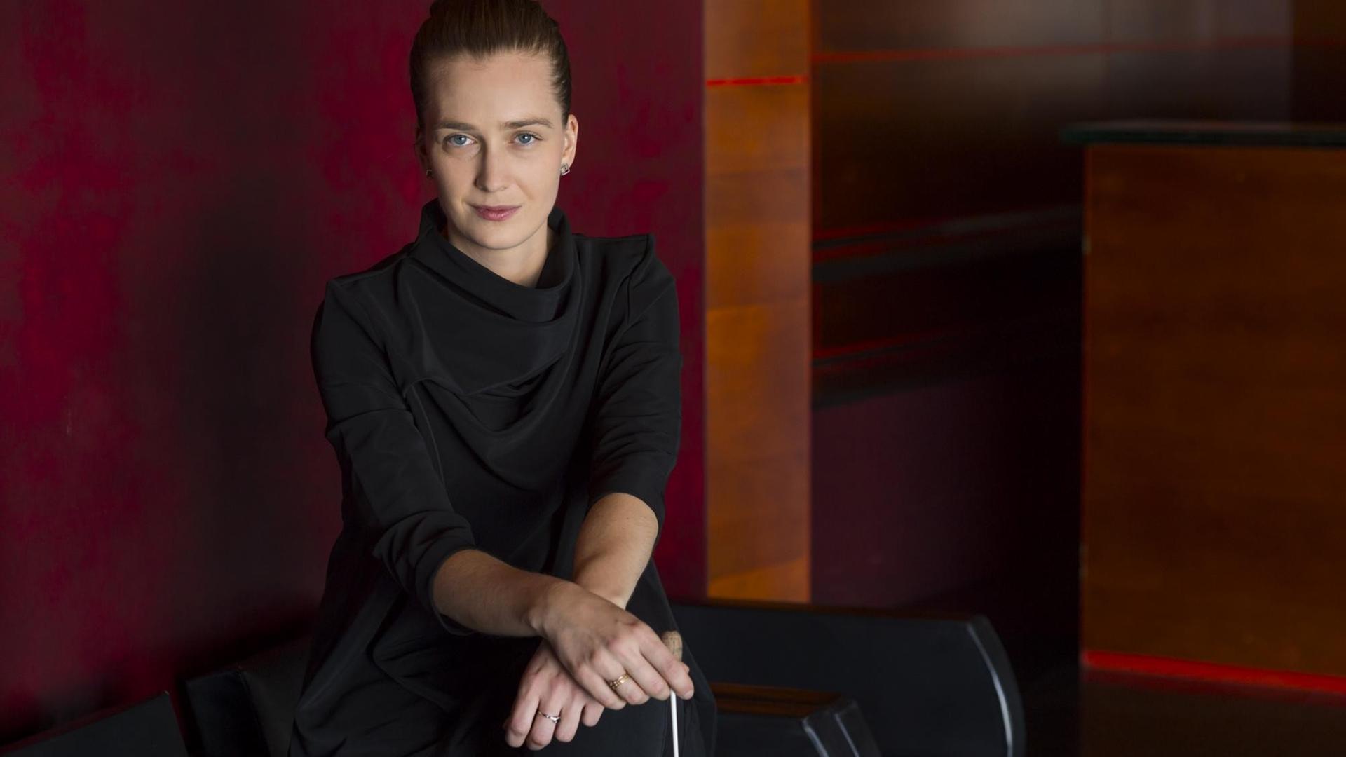 Die junge Dirigentin Elena Schwarz steht in einem schwarzen Shirt mit elegantem Faltenwurf vor einer rötlichen Wand, wobei sie ihre Hände auf einer Stuhllehne über Kreuz gelegt hat und dabei einen Dirigentenstab hält. Die junge Dirigentin Elena Schwarz steht in einem schwarzen Shirt mit elegantem Faltenwurf vor einer rötlichen Wand, wobei sie ihre Hände auf einer Stuhllehne über Kreuz gelegt hat und dabei einen Dirigentenstab hält.
