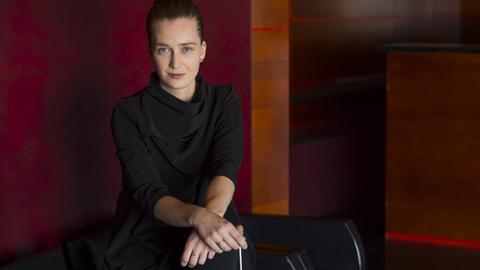 Die junge Dirigentin Elena Schwarz steht in einem schwarzen Shirt mit elegantem Faltenwurf vor einer rötlichen Wand, wobei sie ihre Hände auf einer Stuhllehne über Kreuz gelegt hat und dabei einen Dirigentenstab hält.