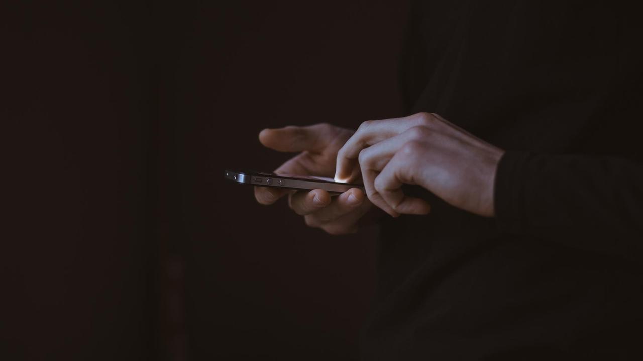 Das Bild zeigt zwei helle Hände vor sehr dunklem Hintergrund, die mit den Fingerspitzen ein leuchtendes Smartphone-Display bedienen. Das Bild zeigt zwei helle Hände vor sehr dunklem Hintergrund, die mit den Fingerspitzen ein leuchtendes Smartphone-Display bedienen.