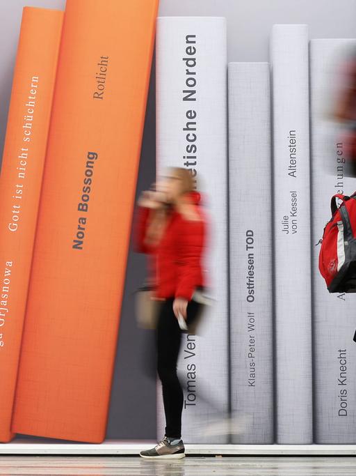 Besucher der Leipziger Buchmesse gehen an einer Fotowand vorbei, auf der überdimensionale Bücher abgebildet sind.