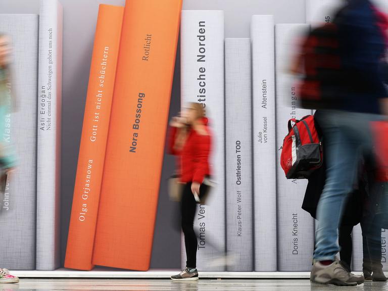 Besucher der Leipziger Buchmesse gehen an einer Fotowand vorbei, auf der überdimensionale Bücher abgebildet sind.