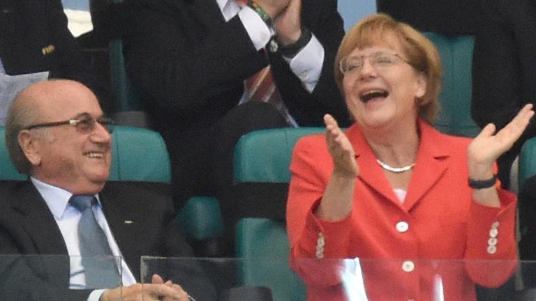 Joseph Blatter sitzt zurückgelehnt lachend und schaut zu Angela Merkel herüber, die lacht und klatscht.