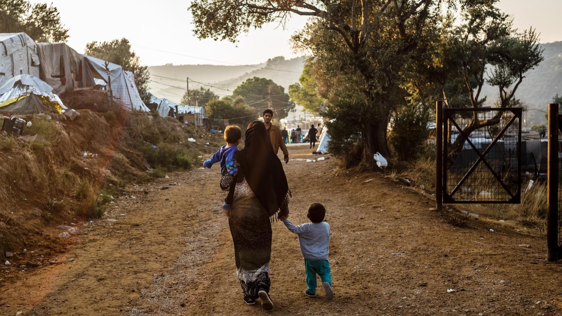 Ein Frau geht mit einem Kind auf dem Arm und einem anderen Kind an der Hand auf einem Weg im Flüchtlingslager Moria auf der griechischen Insel Lesbos, während ihr ein Mann entgegen kommt. Ein Frau geht mit einem Kind auf dem Arm und einem anderen Kind an der Hand auf einem Weg im Flüchtlingslager Moria auf der griechischen Insel Lesbos, während ihr ein Mann entgegen kommt.