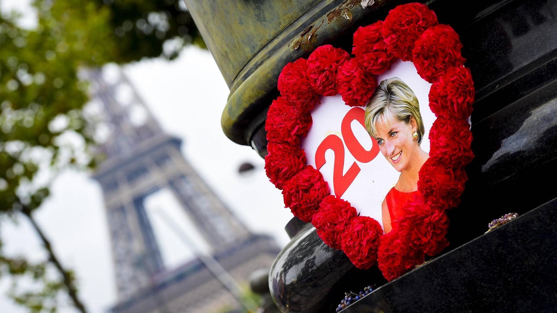 Ein gebasteltes Herz mit rotem Rahmen, in dessen Mitte ein Foto von Prinzessin Diana zu sehen ist, hängt an einer Brücke in Paris. Im Hintergrund ist der Eiffelturm zu erkennen. Ein gebasteltes Herz mit rotem Rahmen, in dessen Mitte ein Foto von Prinzessin Diana zu sehen ist, hängt an einer Brücke in Paris. Im Hintergrund ist der Eiffelturm zu erkennen.