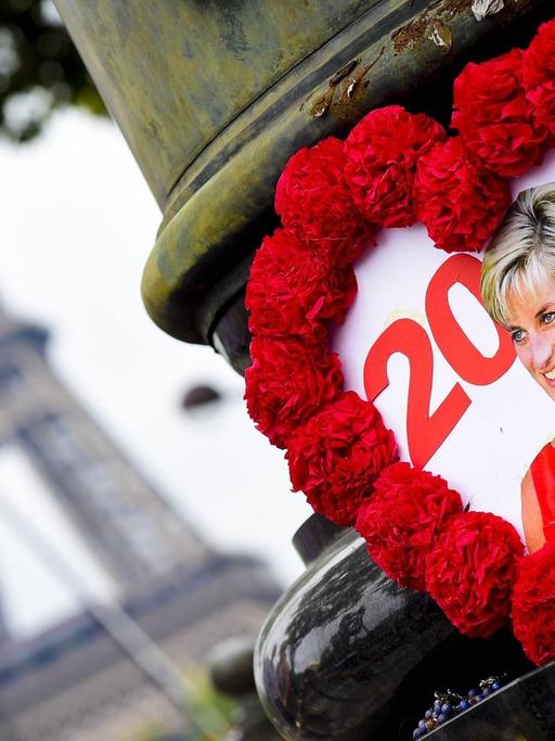 Ein gebasteltes Herz mit rotem Rahmen, in dessen Mitte ein Foto von Prinzessin Diana zu sehen ist, hängt an einer Brücke in Paris. Im Hintergrund ist der Eiffelturm zu erkennen.