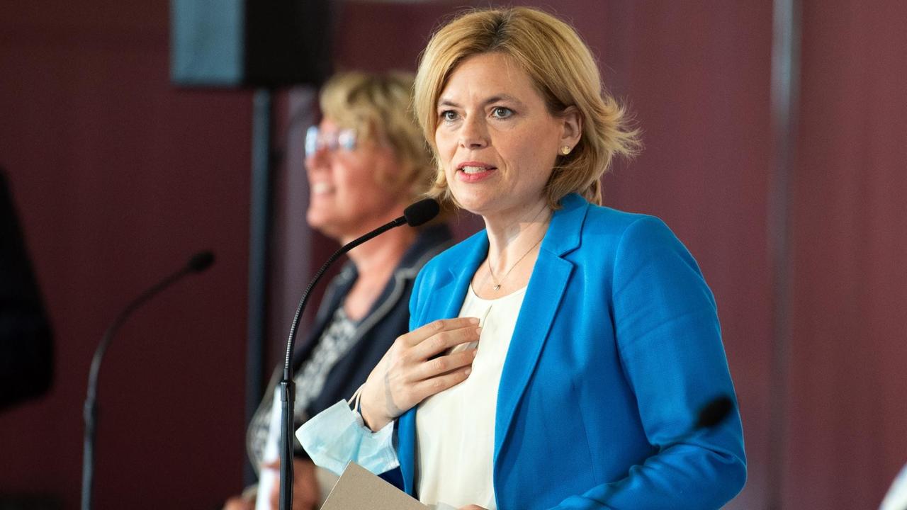 Klöckner steht im blauen Jackett in einem Raum vor einem Mikro und spricht. Sie hält einen Ordner in der Hand. Hinter ihr unscharf eine weitere Frau. Klöckner steht im blauen Jackett in einem Raum vor einem Mikro und spricht. Sie hält einen Ordner in der Hand. Hinter ihr unscharf eine weitere Frau.