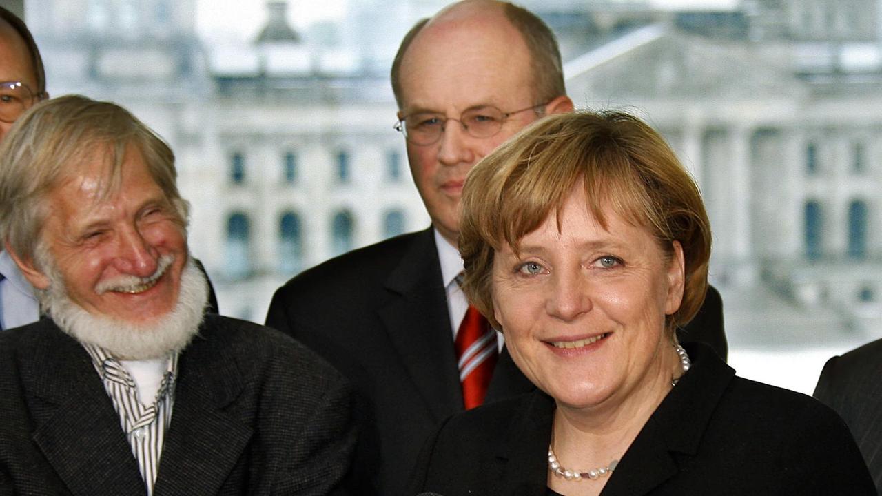Merkel spricht lächelnd in ein Mikrofon, Neudeck steht lachend dahinter neben Kauder. Im Hintergrund sieht man durch ein Fenster den Reichstag.