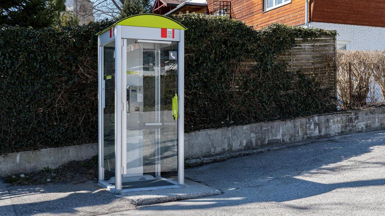 Eine gläserne Telefonzelle steht an einer menschenleeren Straße. Eine gläserne Telefonzelle steht an einer menschenleeren Straße.