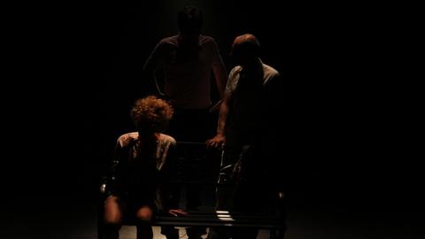 In sehr dunklem Licht sind drei Silhouetten zu erkennen, eine Person sitzt auf einer Bank, die anderen beiden stehen dahinter.