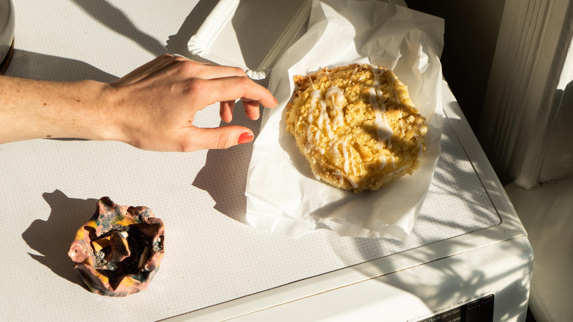 Eine Hand greift nach einem Streuselbrötchen, das neben einem Aschenbecher auf einer Waschmaschine liegt. Eine Hand greift nach einem Streuselbrötchen, das neben einem Aschenbecher auf einer Waschmaschine liegt.