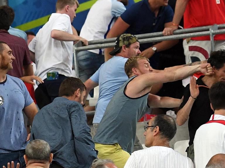 Das Bild zeigt mehrere Hooligans und viele flüchtende Fans; in der Bildmitte schlägt einer der Hooligans einem Zuschauer mit der Faust in den Nacken.