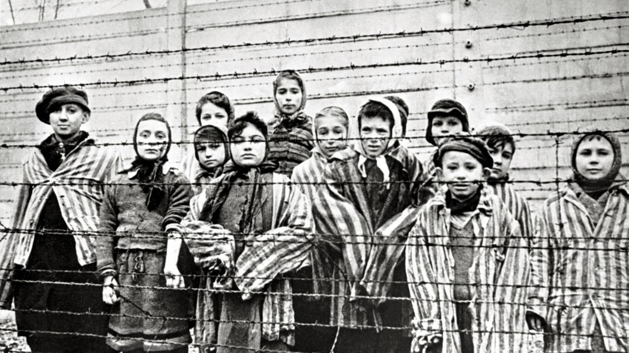 Überlebende jüdische Kinder stehen 1945 in Auschwitz mit einer Krankenschwester hinter Stacheldrahtzaun. Das Foto wurde von einem sowjetischen Fotografen während der Herstellung eines Films über Befreiung des Lagers gemacht. Die Kinder wurden von den Russen mit Kleidung von erwachsenen Gefangenen verkleidet. Überlebende jüdische Kinder stehen 1945 in Auschwitz mit einer Krankenschwester hinter Stacheldrahtzaun. Das Foto wurde von einem sowjetischen Fotografen während der Herstellung eines Films über Befreiung des Lagers gemacht. Die Kinder wurden von den Russen mit Kleidung von erwachsenen Gefangenen verkleidet.