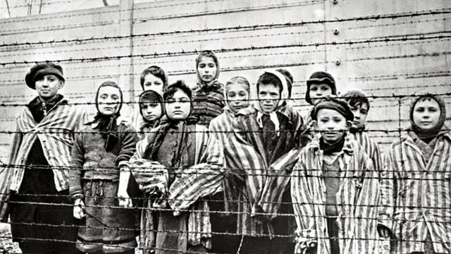 Überlebende jüdische Kinder stehen 1945 in Auschwitz mit einer Krankenschwester hinter Stacheldrahtzaun. Das Foto wurde von einem sowjetischen Fotografen während der Herstellung eines Films über Befreiung des Lagers gemacht. Die Kinder wurden von den Russen mit Kleidung von erwachsenen Gefangenen verkleidet.