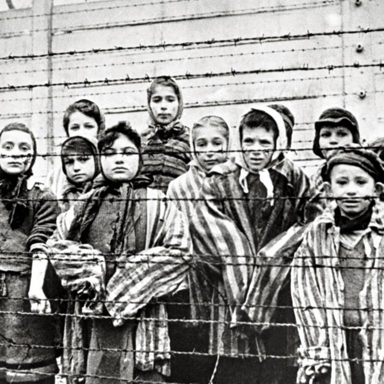 Überlebende jüdische Kinder stehen 1945 in Auschwitz mit einer Krankenschwester hinter Stacheldrahtzaun. Das Foto wurde von einem sowjetischen Fotografen während der Herstellung eines Films über Befreiung des Lagers gemacht. Die Kinder wurden von den Russen mit Kleidung von erwachsenen Gefangenen verkleidet. 