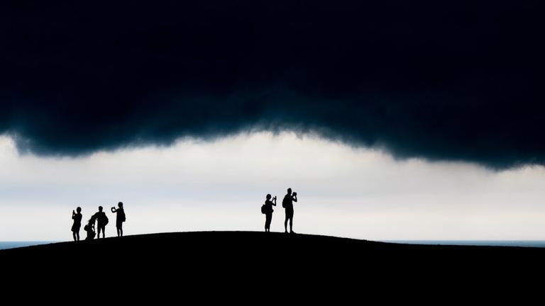Menschen stehen auf einer Anhöhe und machen Bilder von einer tief hängenden schwarzen Wolke Menschen stehen auf einer Anhöhe und machen Bilder von einer tief hängenden schwarzen Wolke