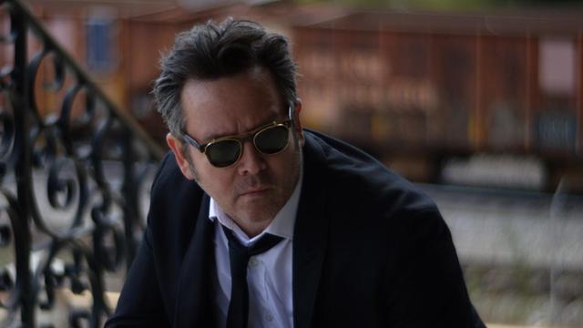 Ein Mann in schwarzem Jackett, weißem Hemd und schwarzer Krawatte sitzt vor einem Treppengeländer und hat eine Sonnenbrille vor den Augen.