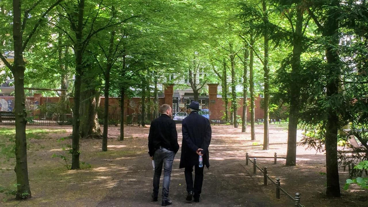 Ilja Richter und Moderator Olaf Kosert gehen durch den Friedhofspark Pappelallee. Das Foto zeigt sie von hinten, wie sie durch eine Allee grüner Bäume auf ein Tor zugehen.