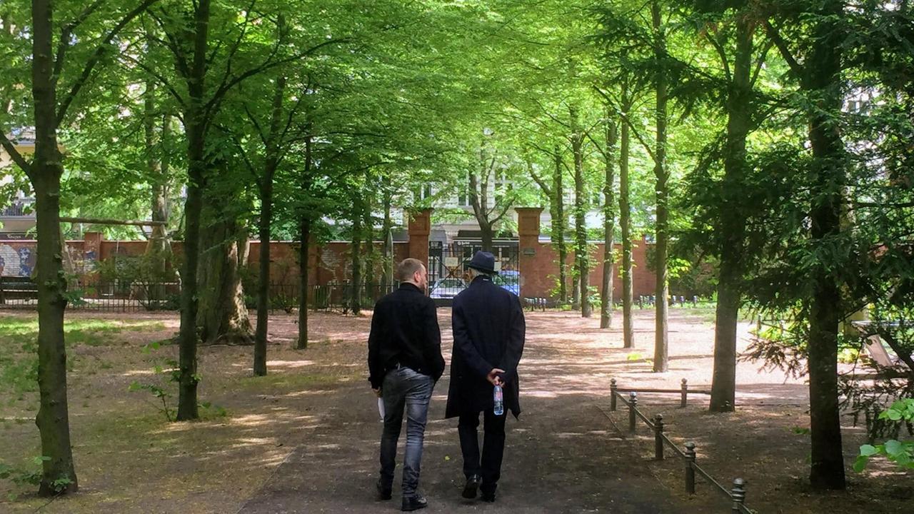 Ilja Richter und Moderator Olaf Kosert gehen durch den Friedhofspark Pappelallee. Das Foto zeigt sie von hinten, wie sie durch eine Allee grüner Bäume auf ein Tor zugehen. Ilja Richter und Moderator Olaf Kosert gehen durch den Friedhofspark Pappelallee. Das Foto zeigt sie von hinten, wie sie durch eine Allee grüner Bäume auf ein Tor zugehen.
