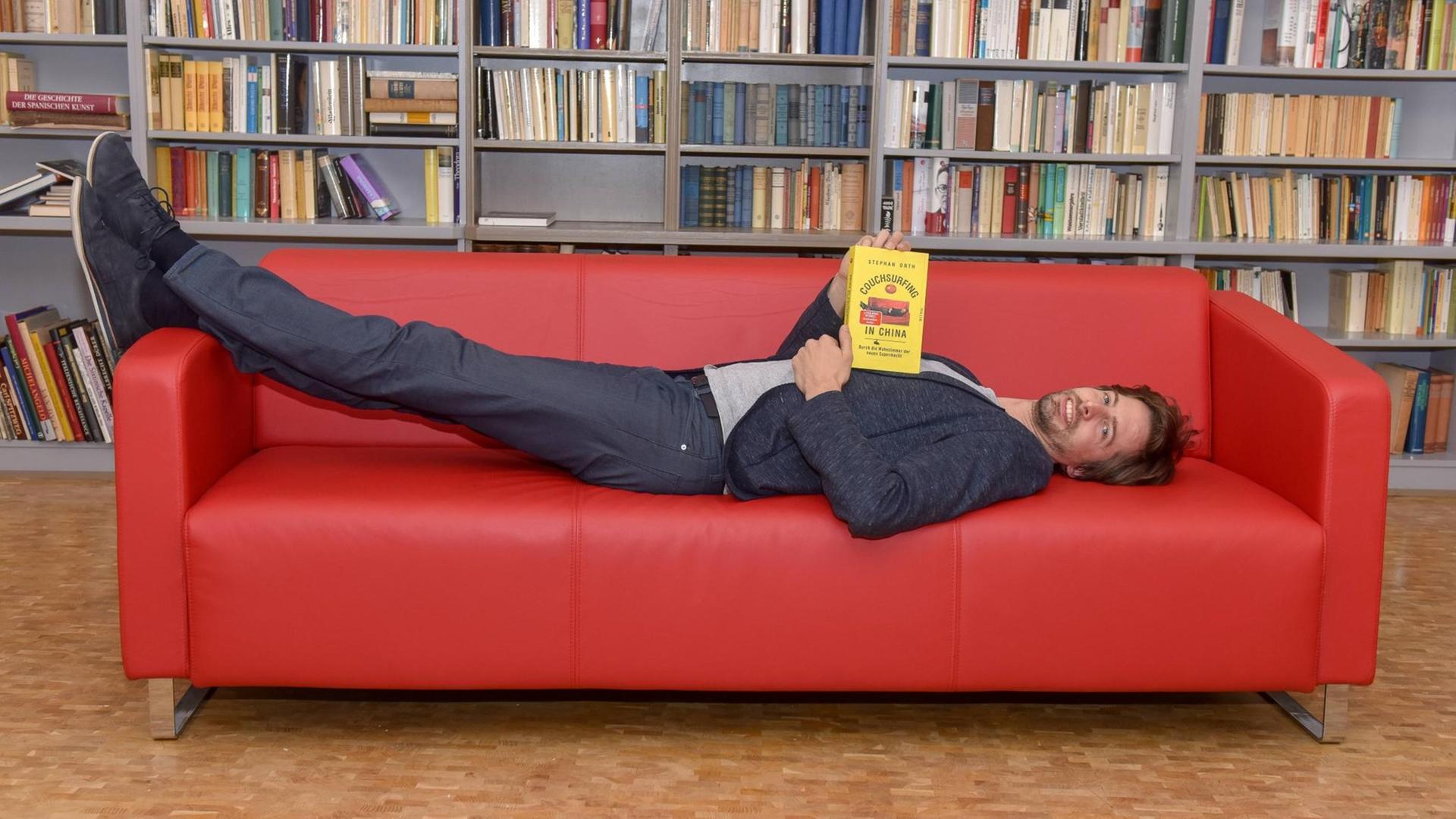 Stephan Orthliegt liegt auf einer roten Couch, einem Möbelstück, welches er auf Reisen als Couchsurfer in China in unterschiedlichen Formen vorgefunden hat. Die Erfahrungen aus dem Couchsurfing sind die Grundlage für sein neues Buch.