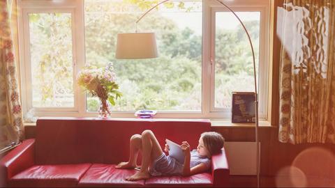 Ein junges Mädchen liegt mit einem Tablet-Computer auf einem roten Sofa. Durchs Fenster sieht man grüne Natur im Hintergrund.