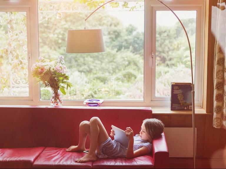 Ein junges Mädchen liegt mit einem Tablet-Computer auf einem roten Sofa. Durchs Fenster sieht man grüne Natur im Hintergrund.