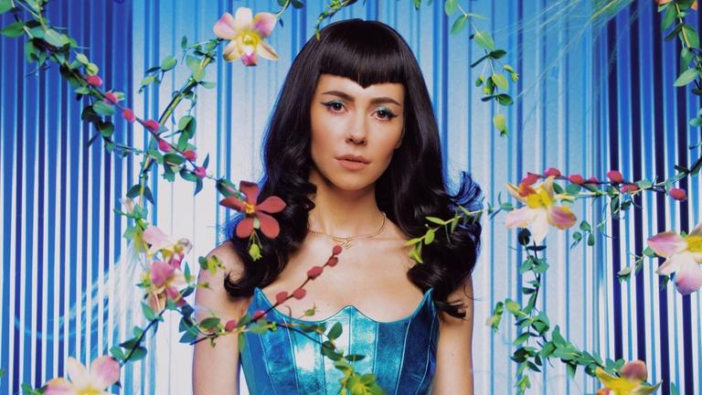 Pressebild zum neuen Album. Die Künstlerin Marina steht vor einer blauen Wand, um sie herum ranken sich Blumenzweige.