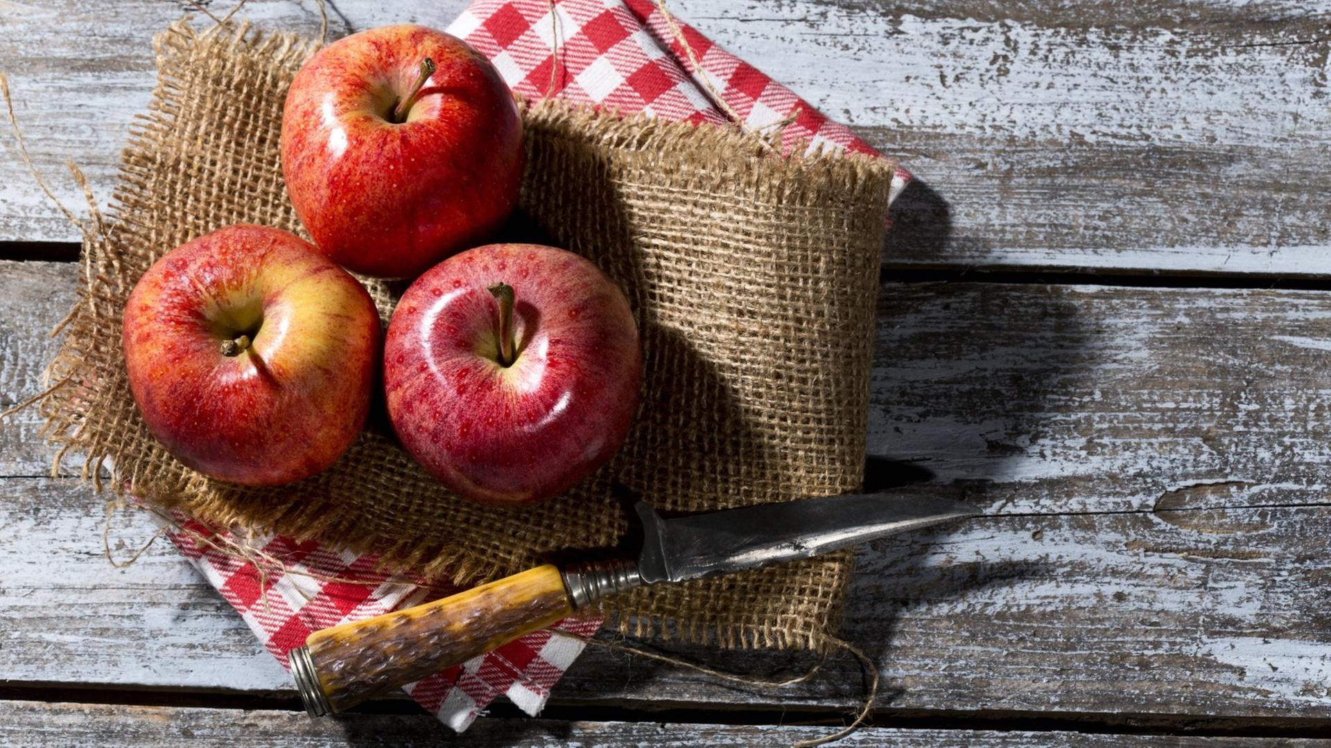 Drei rote Äpfel und ein Messer liegen auf einem Jute-Tuch auf einem Holztisch Drei rote Äpfel und ein Messer liegen auf einem Jute-Tuch auf einem Holztisch