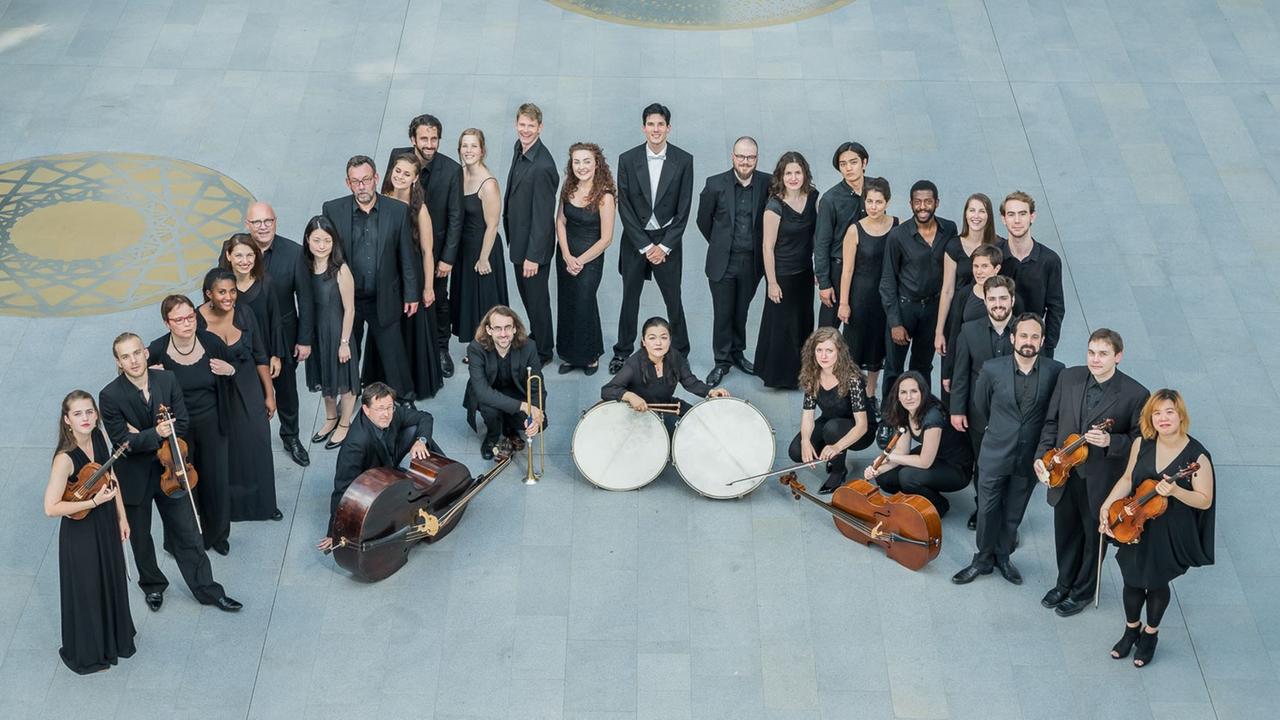 Musiker*innen und Sänger*innen stehen in Konzertkleidung im Halbkreis hinter Pauken, Kontrabass und Cello Musiker*innen und Sänger*innen stehen in Konzertkleidung im Halbkreis hinter Pauken, Kontrabass und Cello