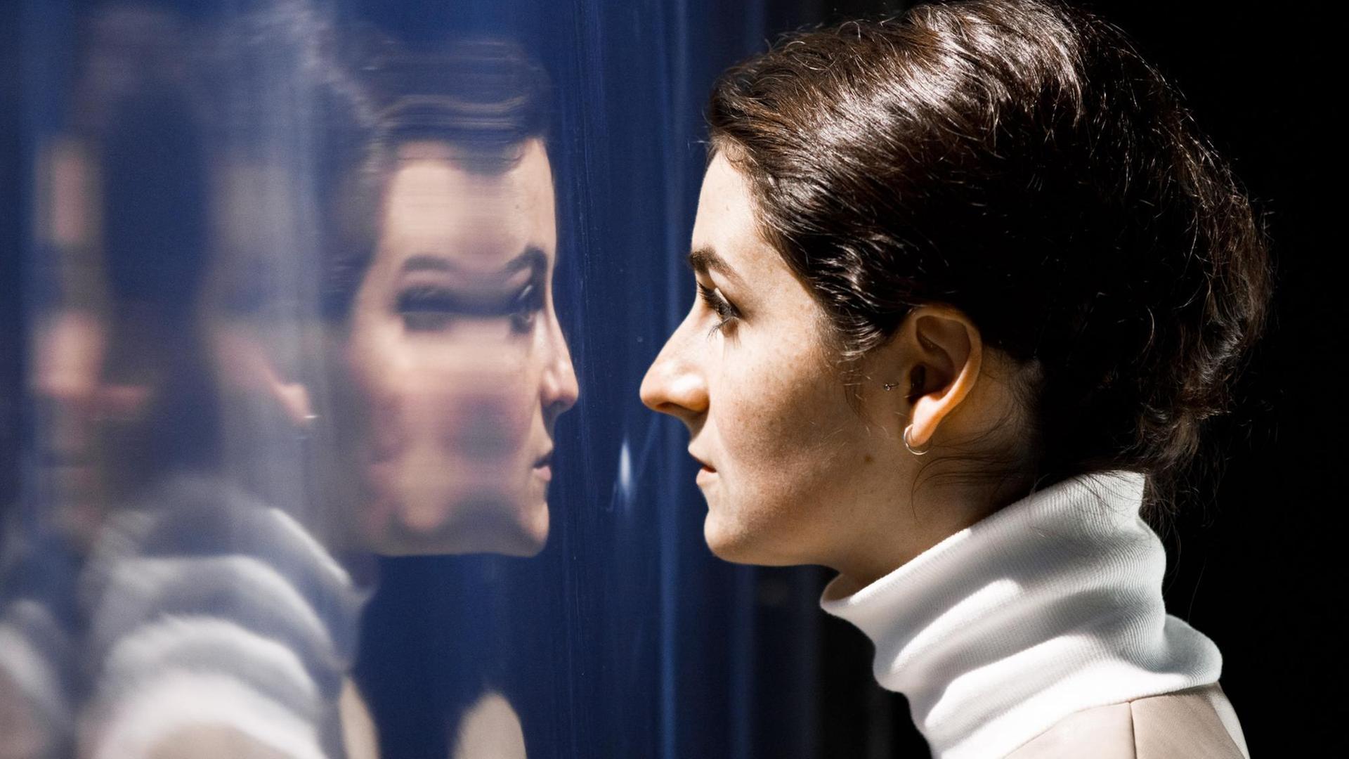 Szene aus der Theaterperformance "How to protect your internal ecosystem": Eine Darstellerin sieht ihre Spiegelung auf einer Trennwand Szene aus der Theaterperformance "How to protect your internal ecosystem": Eine Darstellerin sieht ihre Spiegelung auf einer Trennwand