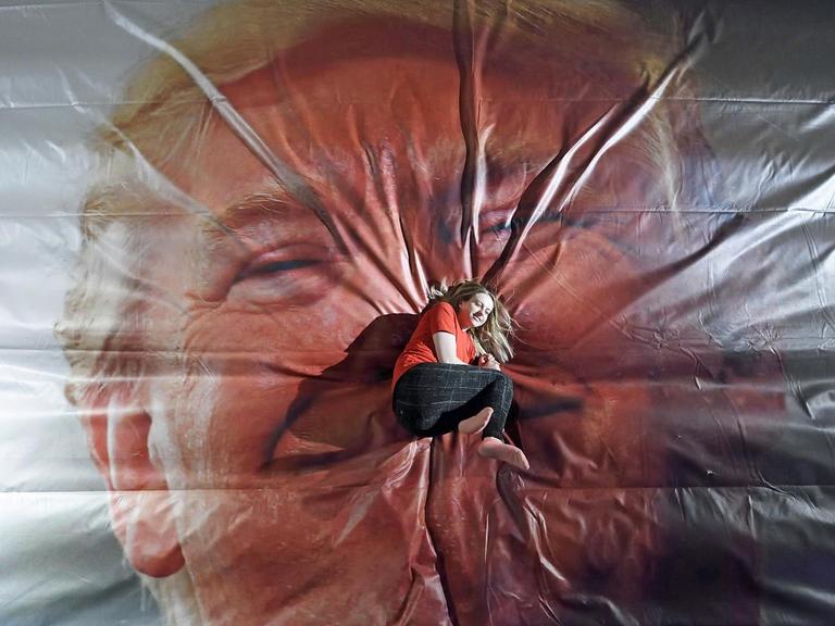 Eine Frau lieget auf einer großen Sprung-Luftmatratze, auf die das Gesicht von Donald Trump aufgedruckt ist.