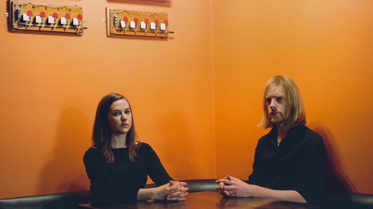 Die Musiker Josienne Clarke und Ben Walker sitzen an einem Tisch vor einer orangenen Wand