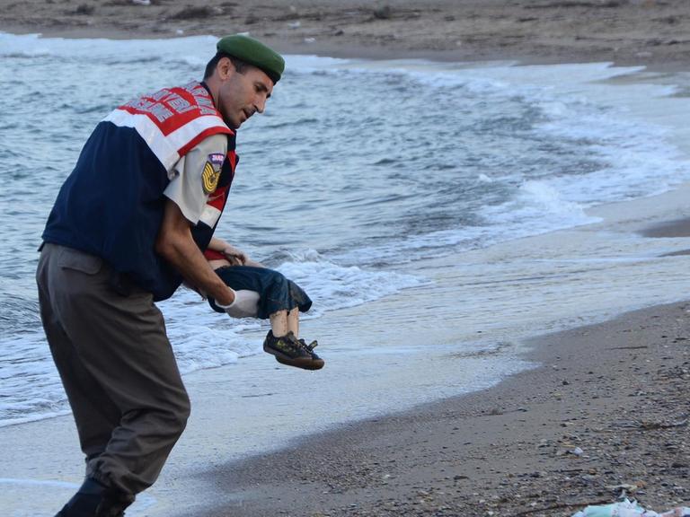 Ein türkischer Polizist trägt ein totes Kind im Arm und bringt es vom Strand weg. Der Oberkörper des Kindes wird vom Körper des Polizisten verdeckt..