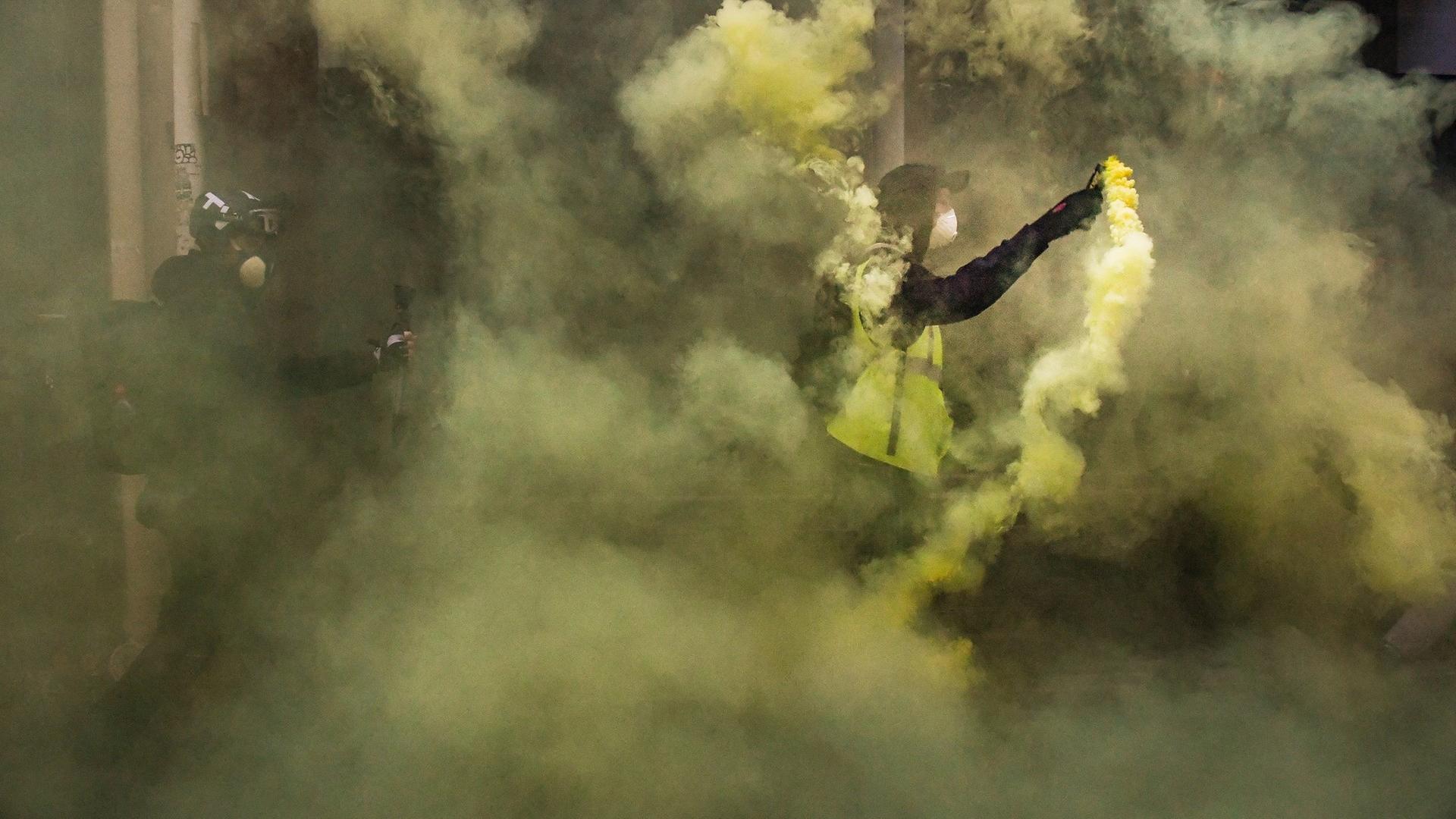 Zwei Männer, einer in einer gelben Warnweste, stehen im gelblichen Rauch von gezündeter Pyrotechnik. Zwei Männer, einer in einer gelben Warnweste, stehen im gelblichen Rauch von gezündeter Pyrotechnik.