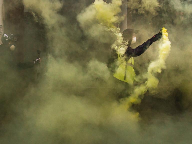 Zwei Männer, einer in einer gelben Warnweste, stehen im gelblichen Rauch von gezündeter Pyrotechnik.