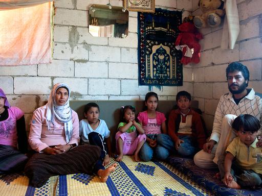 Eine syrische Flüchtlingsfamilie - Mutter, Vater und sechs Kinder - sitzt in Tripolis im Norden des Libanon in einer Wohnung mit unverputzten Wänden auf einem Teppich, an den Wänden hängen ein kleiner Spiegel, einzelne Bilder und zwei große Stoff-Teddybären.