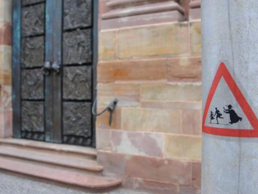 Vor einer Kirchetür im Hintergrund klebt auf einem Pfahl im Vordergrund ein Warnschild. Darauf ein geistlicher, der zwei Kindern hinterher läuft.