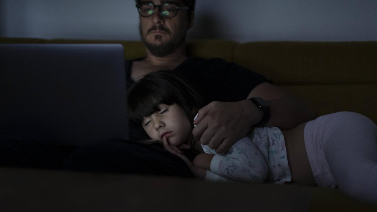 Vater und Tochter sitzen auf der Couch. Der Vater arbeitet am Computer, die Tochter ist auf seinem Schoß eingeschlafen.