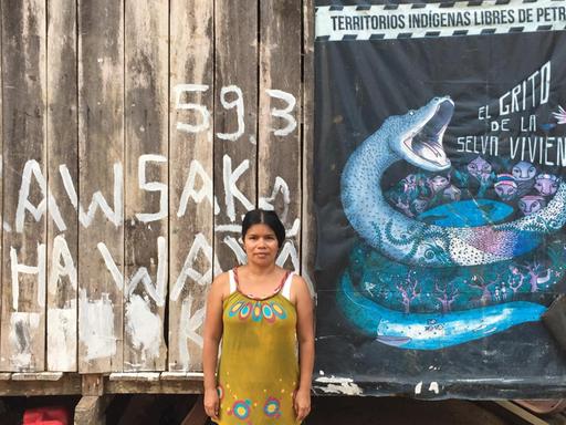Patricia Gualinga ist die Sprecherin der Menschen von Sarayaku. Sie steht vor der Wand eines Holzhauses, an der ein Protestplakat gegen die Erdölförderung in ihrem Lebensraum hängt.