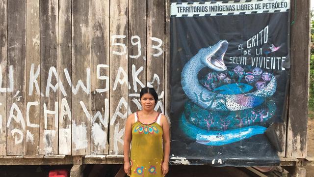 Patricia Gualinga ist die Sprecherin der Menschen von Sarayaku. Sie steht vor der Wand eines Holzhauses, an der ein Protestplakat gegen die Erdölförderung in ihrem Lebensraum hängt.