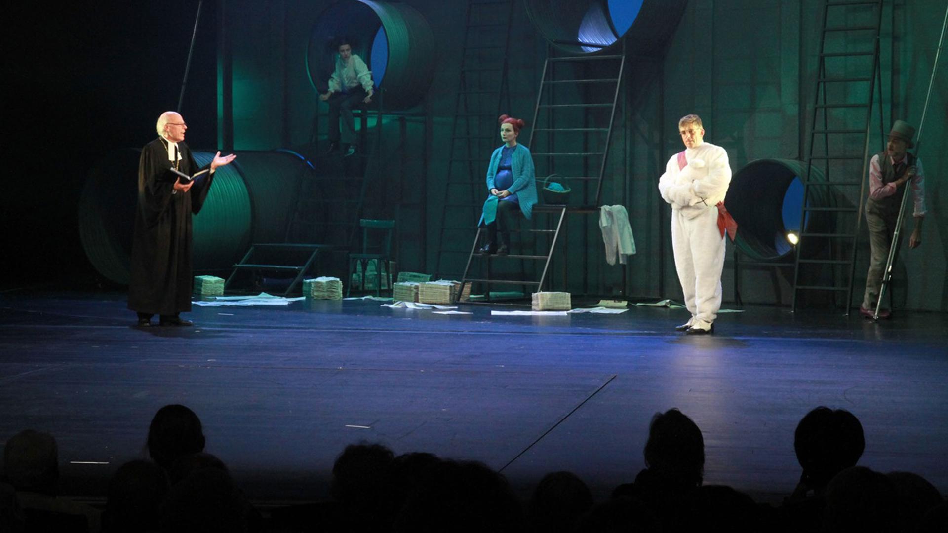 Ein evangelischer Pfarrer steht im Talar und einem Buch in der Hand auf einer Theaterbühne. Auf der in blaues und grünes Licht getauchten Bühne sind vier Schauspieler in Kostümen vor einer Wand mit Bullaugen und Leitern zu sehen.