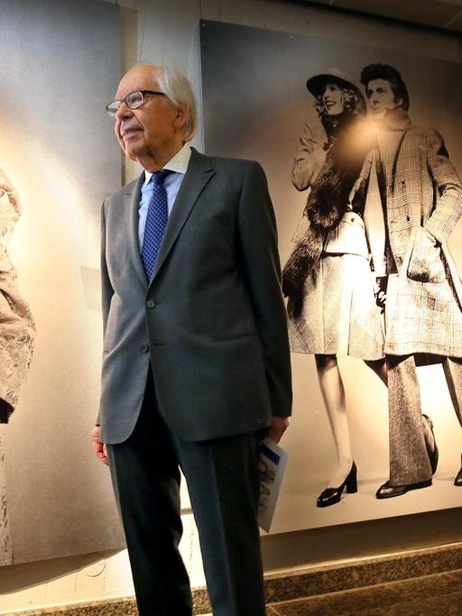 Der Modemacher Uli Richter steht 90-jährig im Anzug vor großen Fotos von Models, die seine Entwürfe targen.