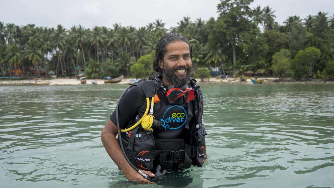 Ein Mann mit schwarzem Vollbart und Tauchausrüstung taucht in Strandnähe aus dem Wasser auf und lächelt.