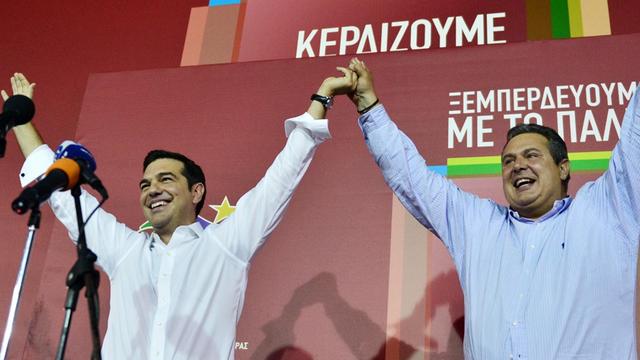 Tsipras und Kamenos reißen auf einer Bühne zusammen die Hände hoch.