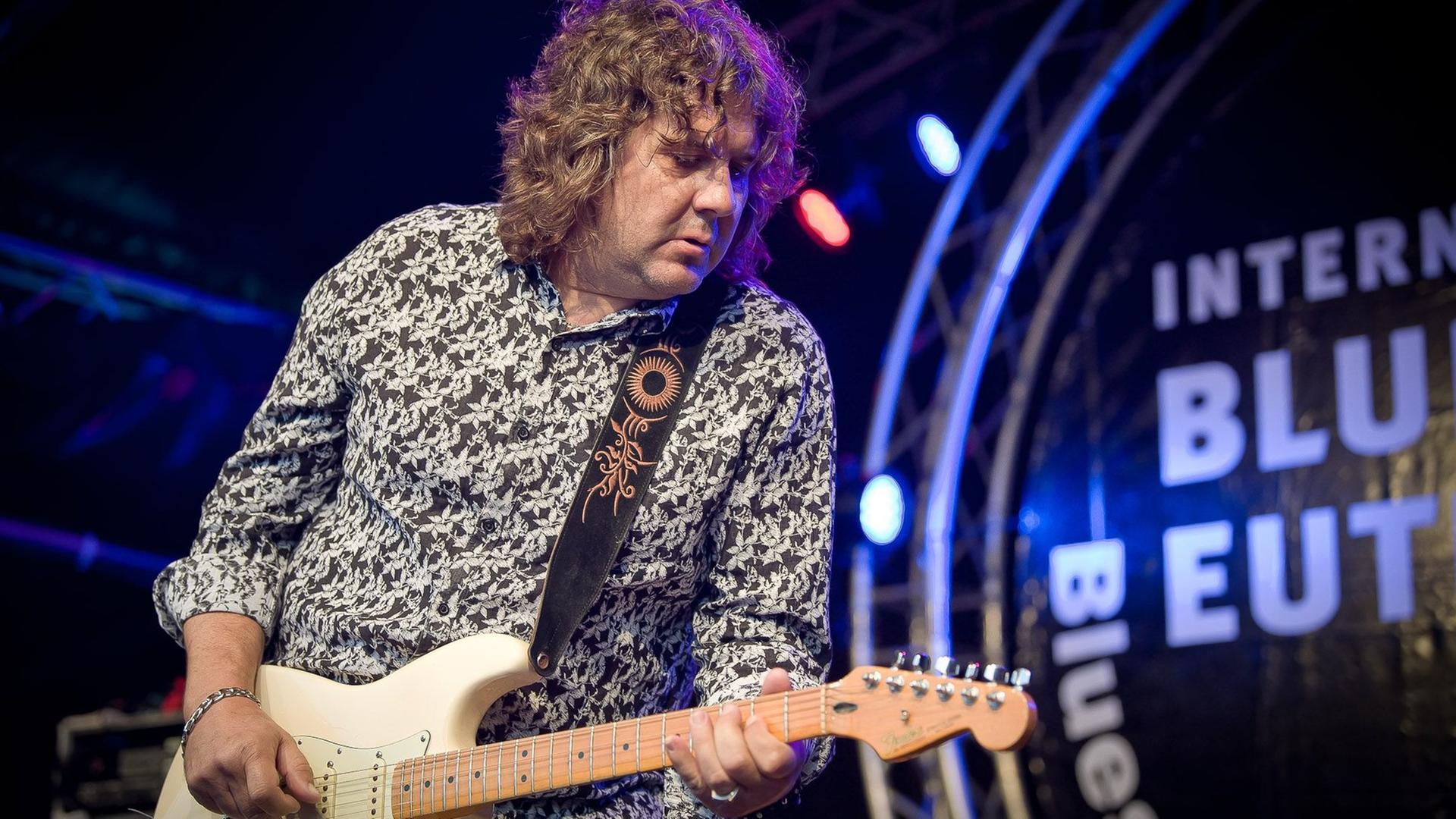 Ein lockiger Mann in buntem Hemd steht auf einer Bühne und spielt E-Gitarre Ein lockiger Mann in buntem Hemd steht auf einer Bühne und spielt E-Gitarre