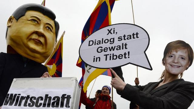 Ein Demonstrant trägt eine Xi-Maske und hält einen Koffer mit der Aufschrift "Wirtschaft" in den Händen. Eine andere trägt eine Merkel-Maske und eine Sprechblase, auf der steht "Xi, Dialog statt Gewalt in Tibet!"