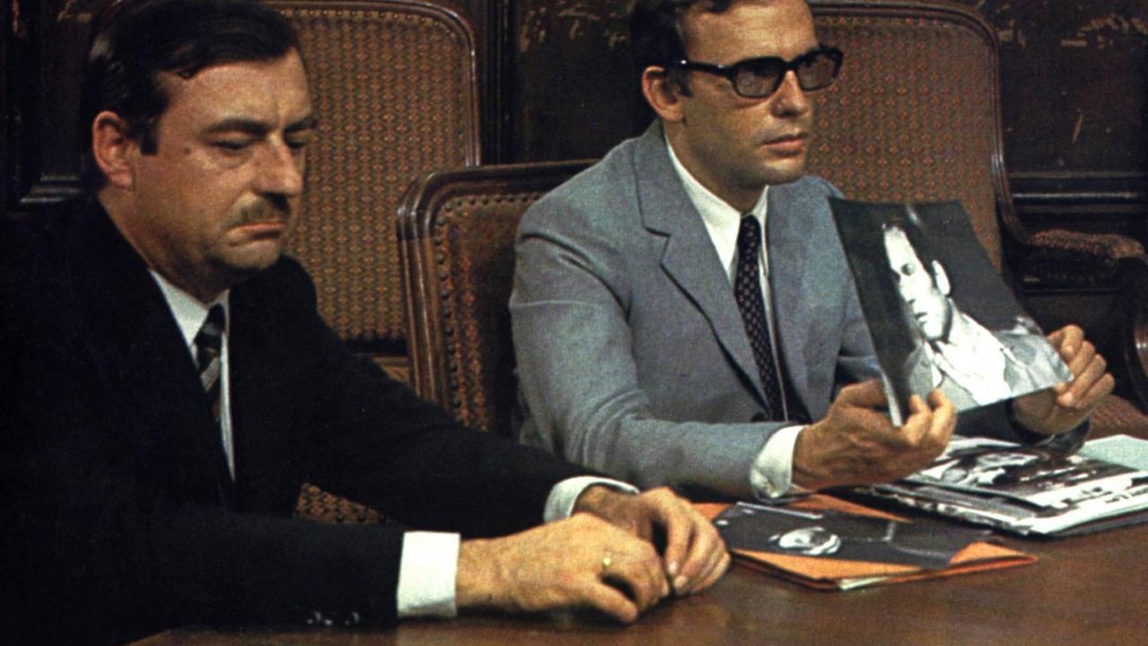Zwei Männer sitzen an einem Konferenztisch, einer von ihnen hält ein Foto hoch.