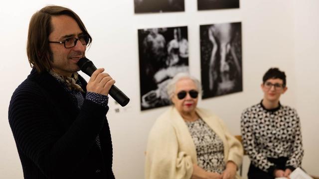Der Regisseur Frank Amann, in ein Mikrofon sprechend, bei der Vernissage zu der Ausstellung "Shot in the dark" in Berlin (mit Sonia Soberats und Moderatorin Kate Brehme im Hintergrund)