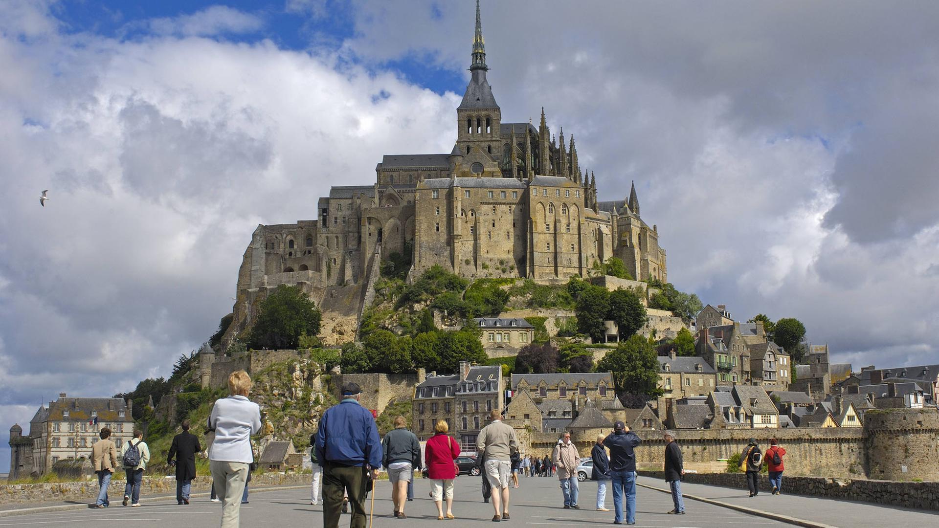 Touristen laufen auf einer Straße auf den Ort Mont-Saint-Michel mit der Abtei in der Normandie in Frankreich zu. Touristen laufen auf einer Straße auf den Ort Mont-Saint-Michel mit der Abtei in der Normandie in Frankreich zu.